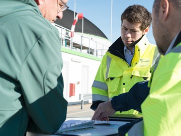 Drei Personen im Gespräch über Unterlagen vor einem Industriegebäude Drei Personen im Gespräch über Unterlagen vor einem Industriegebäude