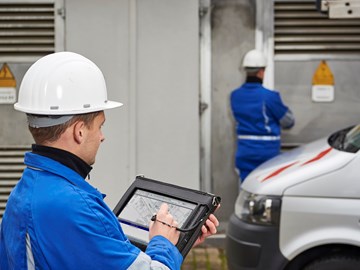 Techniker in Arbeitskleidung mit Eingabegerät, auf dem Pläne zu sehen sind, vor einer Trafostation Techniker in Arbeitskleidung mit Eingabegerät, auf dem Pläne zu sehen sind, vor einer Trafostation
