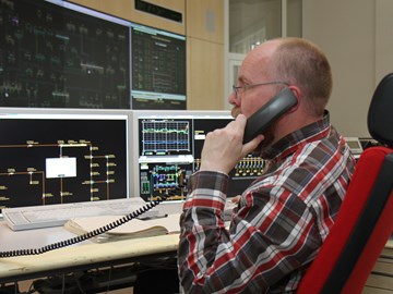 Mitarbeiter telefoniert am Schreibtisch vor Monitoren der Schaltleitung Mitarbeiter telefoniert am Schreibtisch vor Monitoren der Schaltleitung