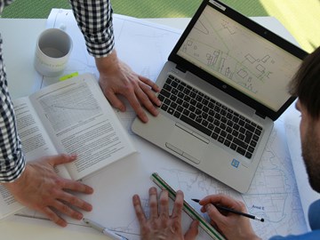 Tisch mit Laptop, Plänen, offenem Buch und Händen zweier Männer - Sicht von oben Tisch mit Laptop, Plänen, offenem Buch und Händen zweier Männer - Sicht von oben